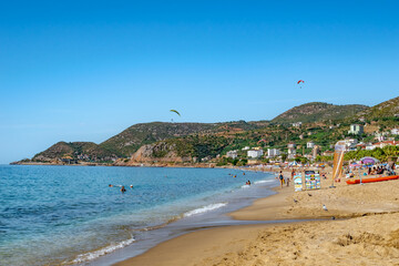 Alanya, Turkey - October 23, 2020: Beautiful summer-autumn view of Kleopatra beach in Alanya. Coastline with clean yellow sand, blue-turquoise water and people resting on the backdrop of mountains - obrazy, fototapety, plakaty
