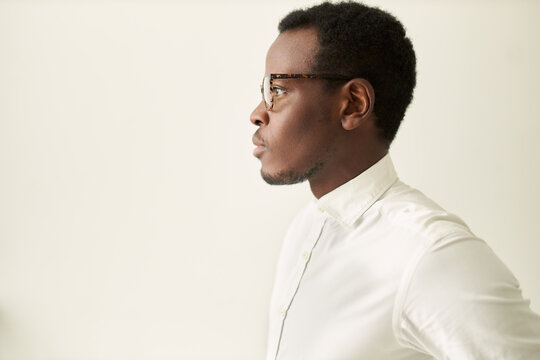 Profile image of attractive serious young dark skinned manager in spectacles having thoughtful pensive facial expression posing against white studio wall background with copy space for your text