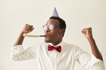 Happy overjoyed young dark skinned man wearing bow tie and cone hat on his head raising clenched...