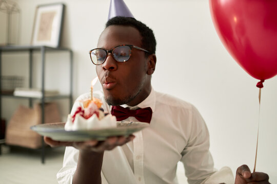 Attractive dreamy young dark skinned man with beard posing indoors with festive decorations wearing glasses and cone hat keeping eyes closed, pouting lips while blowing out candle on cake, making wish