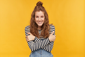 Pleased young ginger female wears denim overalls and stripped shirt, looks into camera with happy facial expression, smiles broadly and hug herself. isolated over yellow background.