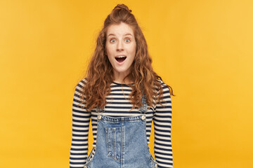 Fototapeta premium indoor portrait of amazed young ginger girl, wears denim overalls and stripped shirt, starring into camera with widely opened mouth and eyes. shocked, amazed facial expression.