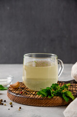 Chicken broth in a glass cup with parsley, garlic and other spices on a wooden board on a gray concrete background. Copy space.