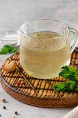 Chicken broth in a glass cup with parsley, garlic and other spices on a wooden board on a gray concrete background. Copy space.