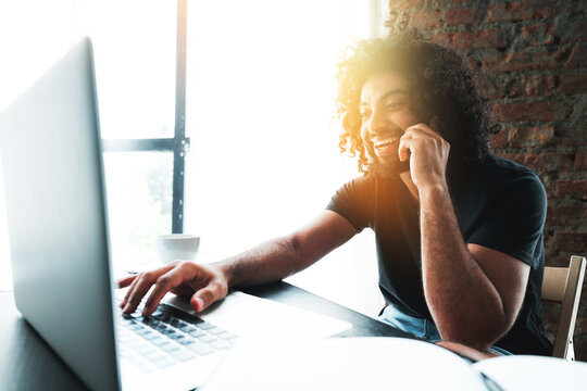  a curly-haired man talking on the phone in a loft cafe and working at a laptop. videoconference concept