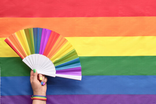 Hand with hand fan on rainbow LGBTQIA background, flat lay with text space. Simple, minimal LGBT pride decor for June, awareness month. Hand with fake painted nails and wrist ribbon.