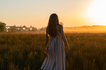 a girl in a dress walks through a field with fog at sunset towards the sun.