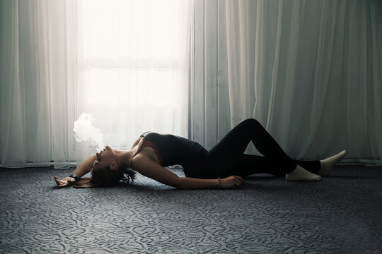 Silhouette of pretty young woman lying and smoking on the floor near big window