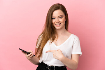 Naklejka premium Teenager girl over isolated pink background using mobile phone and pointing it