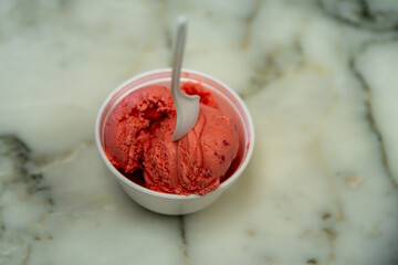 Overhead view of white plastic single-use spoon in expanded polystyrene bowl full of strawberry ice cream scoops on marble table top. 