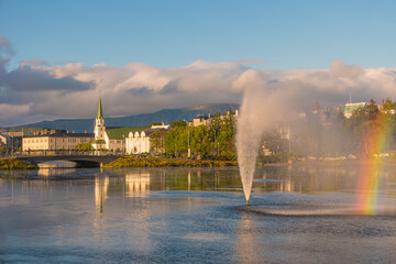 Beautiful view over historical downtown in Reykjavik at warm Summer sunset in the city lake Tjornin with rainbow and fountain, and city park, Iceland.