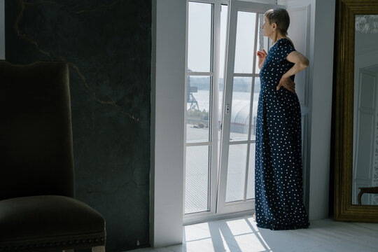 Beautiful elderly woman wearing dress standing and looking out of large window