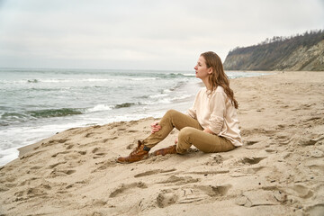 Fototapeta na wymiar A beautiful woman sits on the sand on the beach and looks into the distance of the sea at the ocean horizon. Fall clothes vacation. Caucasian girl is happy Beige clothes and sand in the fall.