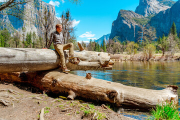 A young man sits on the bank of a river and enjoys the view of a mountain waterfall in Yosemite National Park