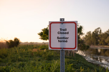 Sign  trail closed in English and French at the park entrance at sunset time. Closure of outdoor facilities, national forests and provincial parks during novel corona virus outbreak quarantine.