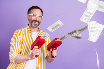 Photo of confident lucky mature man dressed striped shirt spectacles smiling shooting money gun isolated violet color background