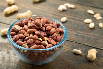 Peeled peanuts in a glass bowl and unpeeled on a wooden table.