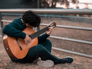 boy playing classical guitar sitting on the ground in a square in autumn in casual clothes