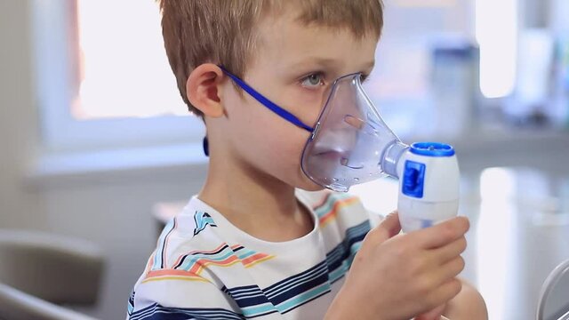 a five-year-old boy in a blue striped T-shirt does inhalation. Allergies, colds, coughs, respiratory diseases, nebulizer. In the video, the child is holding a nebulizer mask and looking at the camera 