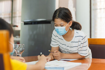 Asian sister in protective mask teaching younger brother homework.