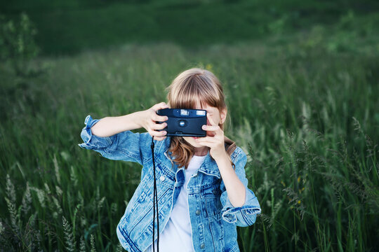 Girl holds camera, hiding among grass in meadow. Child with old film camera takes pictures on summer day in flower field. Childhood, enjoy life, learn photography concept. Copy space