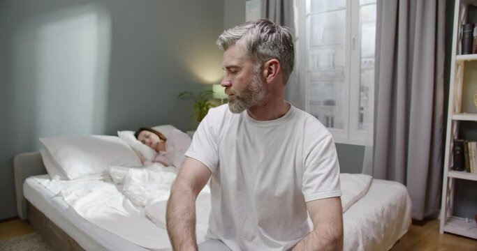 So upset. Waist up portrait view of the sad bearded man sitting at the bed and looking at his sleeping wife after the quarrel. Family disagreements concept