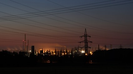 Night photo of a manufacturing plant. High voltage poles in the foreground. Crossing of high voltage wires.