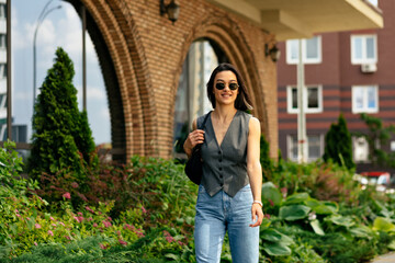 Positive woman in grey vest and jeans walking down the street with bag. Stylish girl with handbag in sunglasses outdoor