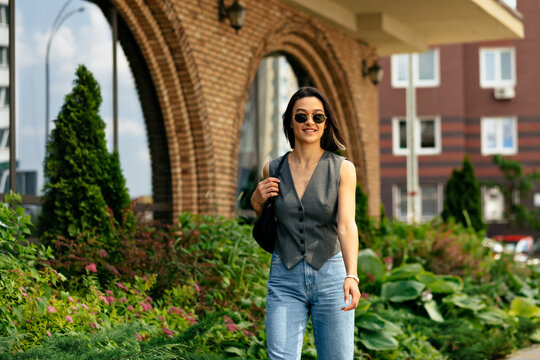 Positive Woman In Grey Vest And Jeans Walking Down The Street With Bag. Stylish Girl With Handbag In Sunglasses Outdoor