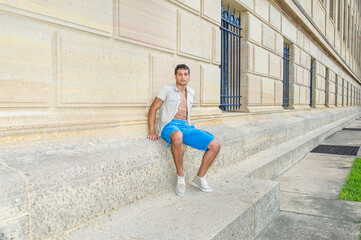 Dressing in a iron short sleeve shirt, blue shorts and sneakers , a young attractive guy is sitting against a pattern wall and relaxing.