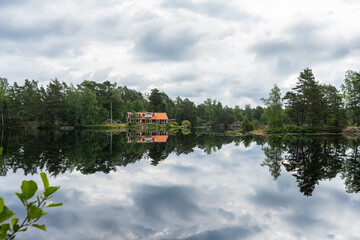 Reflections in the water. Bay lake surface with beautiful red wooden house in the forest among the trees. Green woods pines trees are reflected in the water. Summer forest landscape. Cloudy sky.
