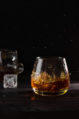 A glass of cognac with ice, splash, a decanter in the background. Alcoholic beverages, dark...