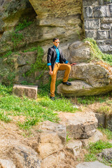 Dressing in a black blazer, a blue shirt, brown pants and a black bow tie,  a young businessman wearing sunglasses is standing on rocks and watching.