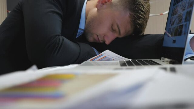 Exhausted boss in formal suit falling asleep on table at boardroom. Tired manager sleeping on table with laptops and financial reports at office. Young entrepreneur napping at workplace. Close up