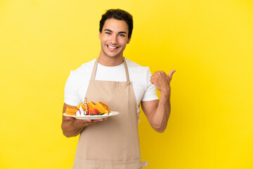 Restaurant waiter holding waffles over isolated yellow background pointing to the side to present a product