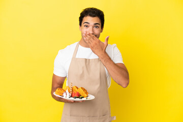 Restaurant waiter holding waffles over isolated yellow background happy and smiling covering mouth with hand