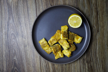 Fried hake fish in a dark plate on a wooden table background. View from above.