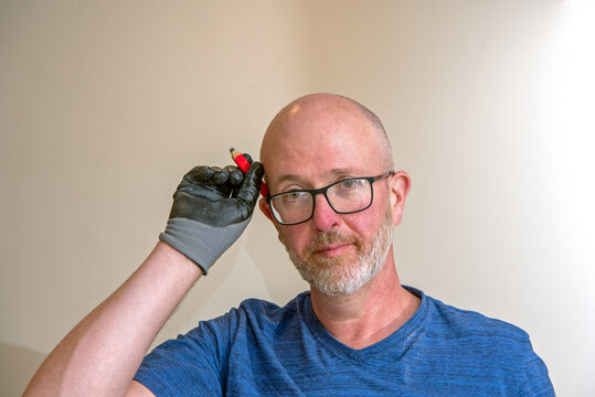 Gay Middle Aged White Man With Glasses And Trimmed Grey Beard Pulls A Carpenter's Pencil From Behind His Ear Using A Gloved Hand.