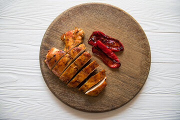 Baked chicken fillet and baked red peppers on a wooden cutting board against a white wooden table background. View from above.