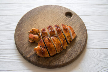 Baked chicken fillet on a wooden cutting board on the background of a white wooden table with a knife and fork.