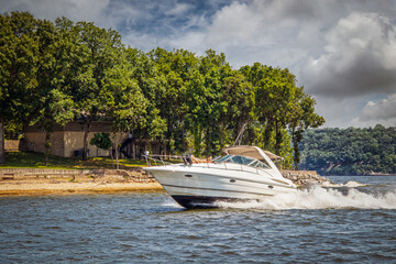 Fototapeta premium Speedboat zooming through water around point at lake with girls suntanning on bow at golden hour under partyly cloudy skies
