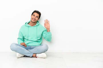 Caucasian handsome man sitting on the floor saluting with hand with happy expression