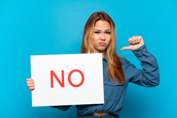 Teenager girl over isolated blue background holding a placard with text NO and doing bad signal