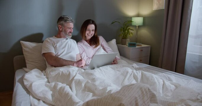 Image of romantic cheerful couple using laptop and hugging while lying on the bed in night room at home. Man bonding to his wife while she is looking at the screen and telling something