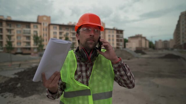 Confident man in protective overalls talking on mobile phone at construction site. Frustrated worker with blueprints and construction plan use cell phone angrily. Supervisor with papers angry.