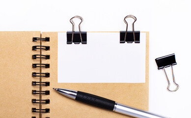 On a light background, a brown notebook with a pen, black clips and a white card with a place to insert text or illustrations. Template.