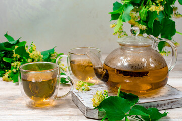 A glass teapot and two cups of tea with a linden tree on a wooden table against a background of linden leaves and flowers. Healing tea.