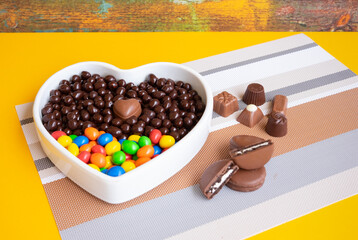 White heart-shaped bowl filled with brown and colorful chocolates, hazelnuts and almonds covered in...
