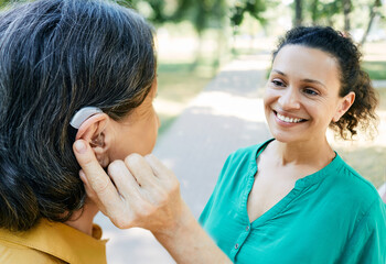Mature woman with a hearing impairment uses a hearing aid to communicate with her female friend...