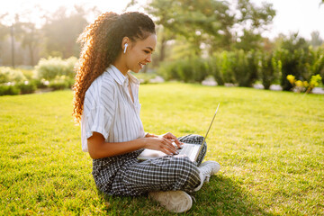 Freelancer woman using laptop computer sitting on grass at park. Young woman working online or...